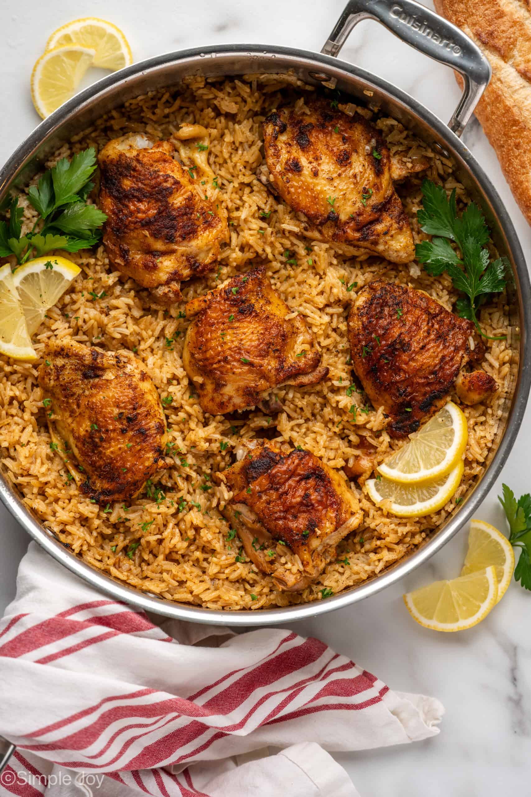 Overhead of skillet Chicken and Rice, garnished with lemon slices and parsley. Lemons slices, parsley, and bread sitting beside.