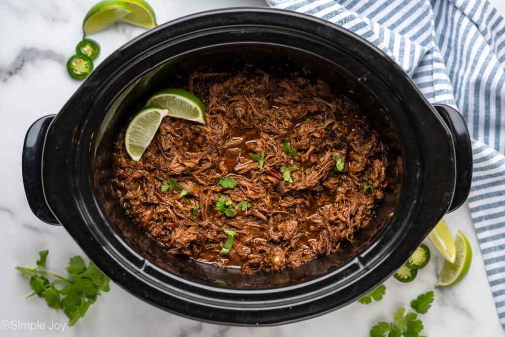 Overhead of beef barbacoa recipe topped with cilantro and limes, diced jalapenos, lime wedges, and cilantro sitting beside