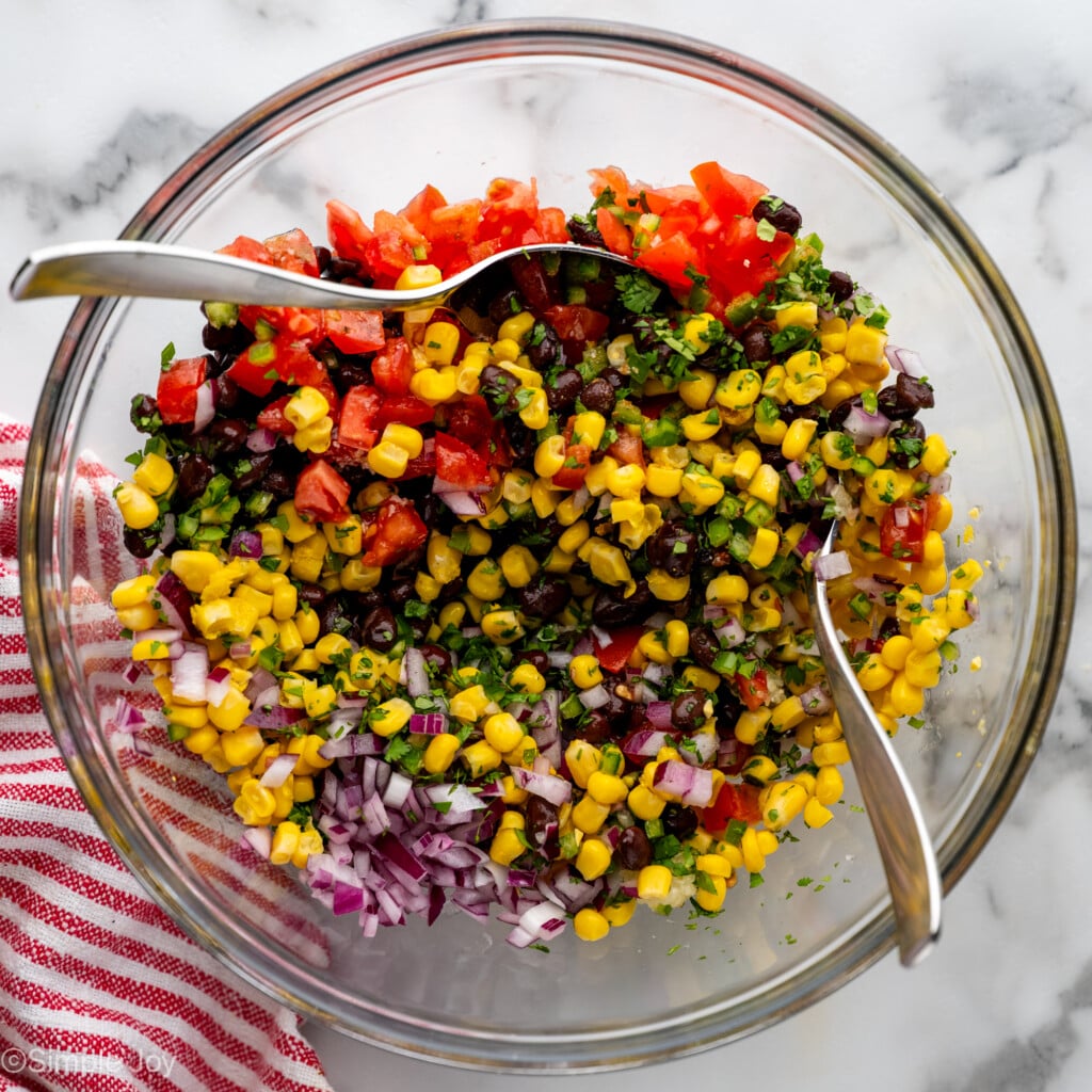overhead of glass bowl of Black Bean and Corn Salsa ingredients with two spoons mixing
