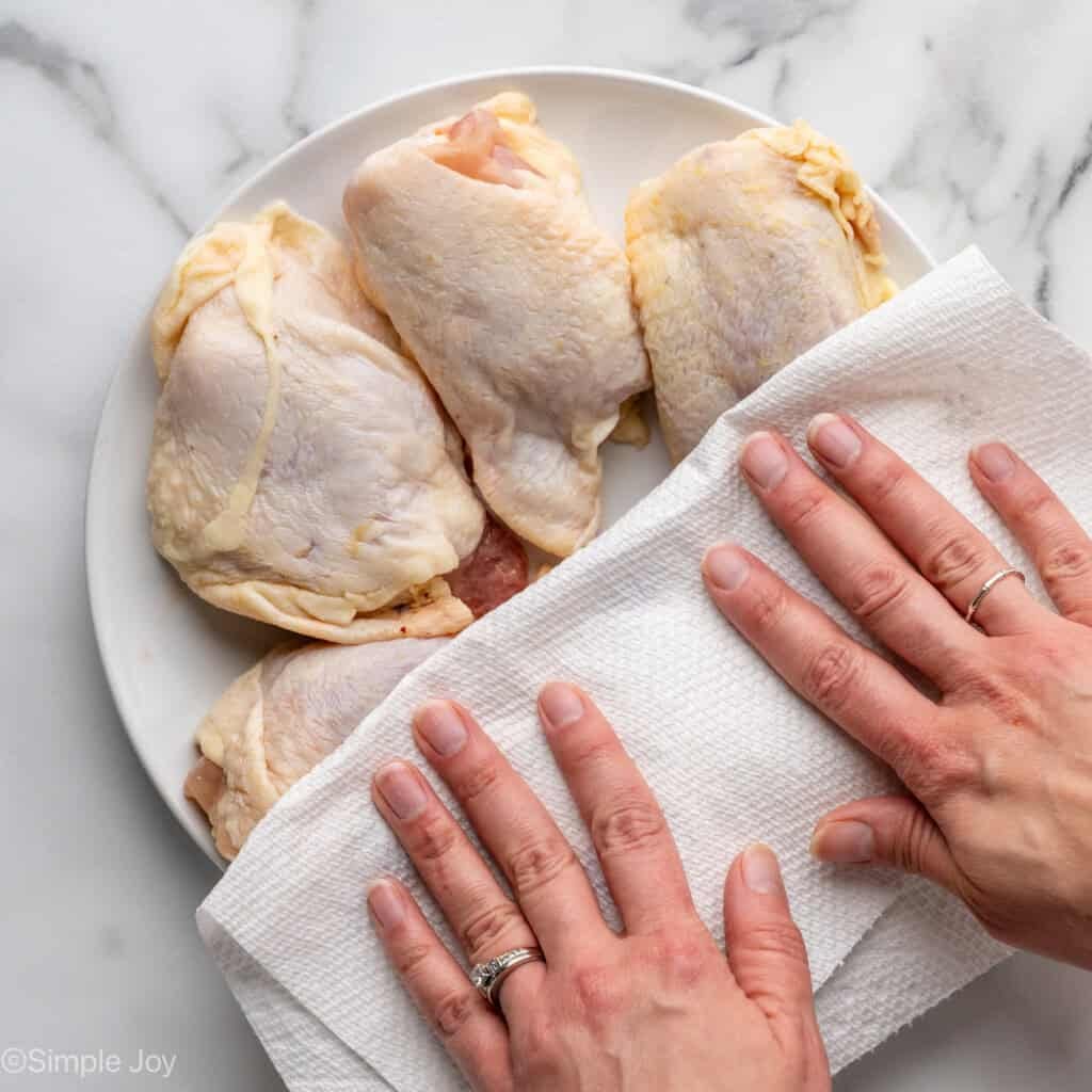 Overhead of person's hand pressing paper towel onto plate of chicken thighs for Chicken and Rice recipe.