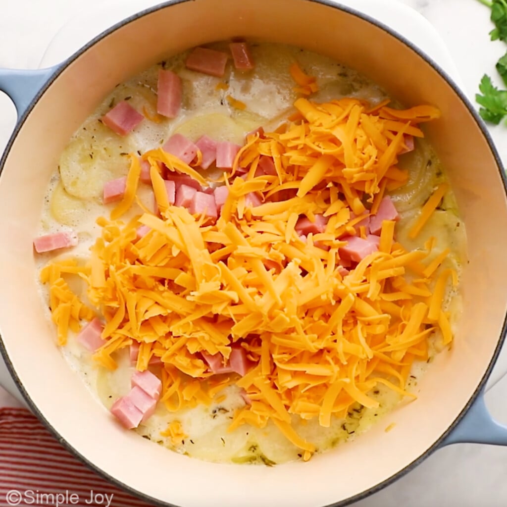 overhead of Dutch oven of scalloped potatoes and ham ingredients with shredded cheese