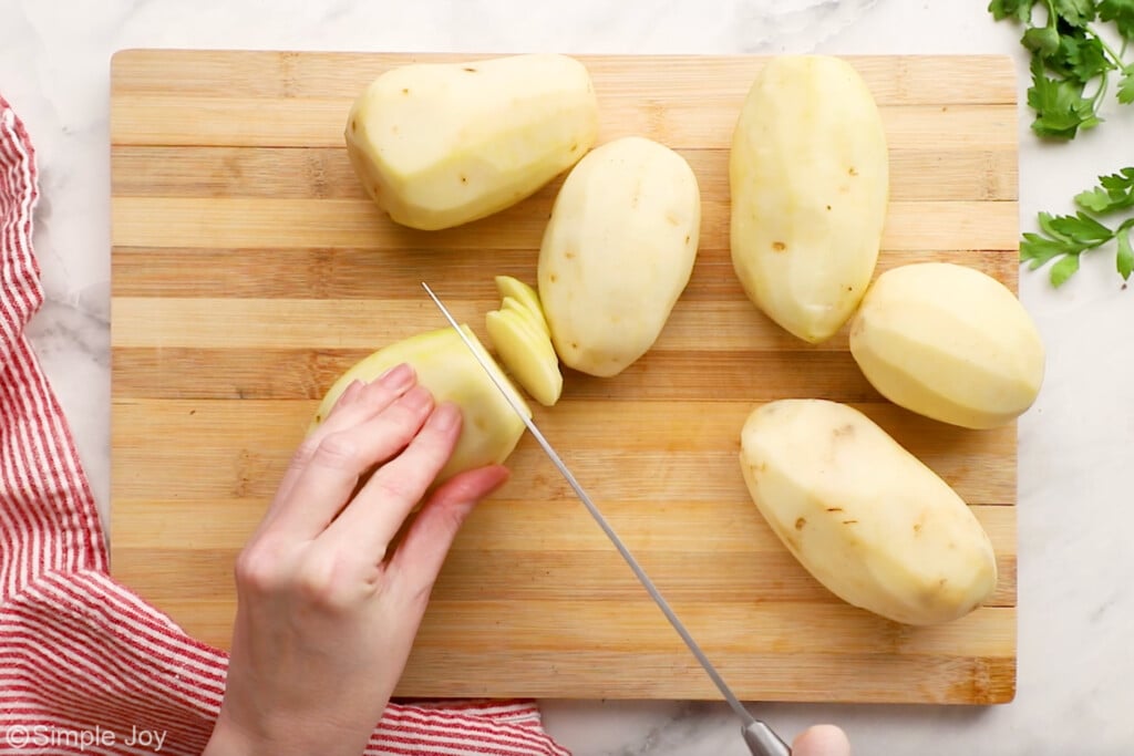 overhead of wooden cutting board with person's hands slicing potato with kitchen knife, with peeled potatoes sitting beside to make scalloped potatoes and ham recipe