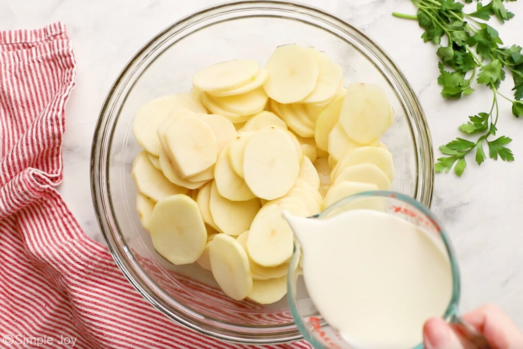 Overhead of glass bowl of sliced potatoes with measuring cup of milk pouring into the bowl to make scalloped potatoes and ham