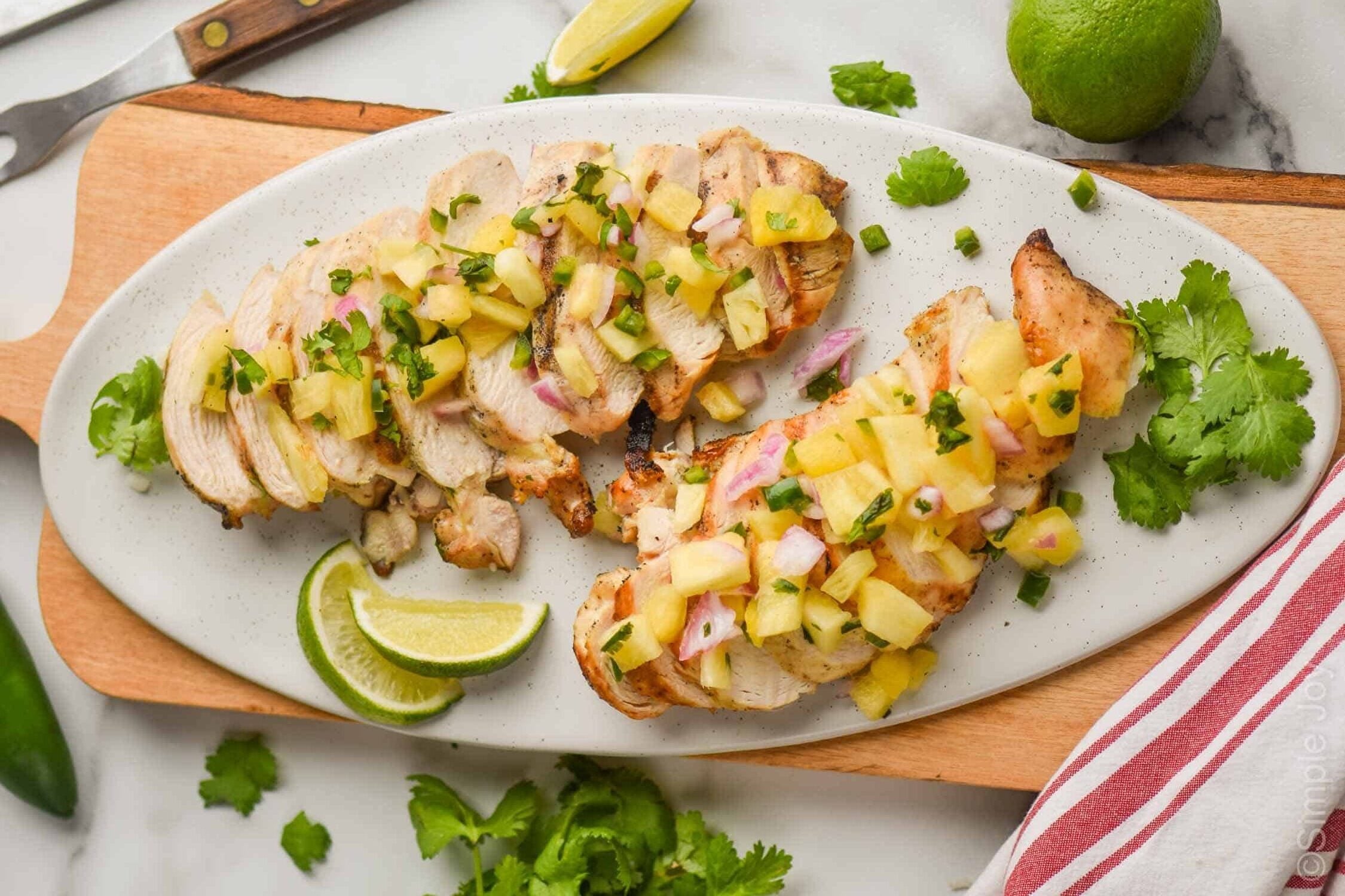 two grilled, sliced chicken breasts covered in pineapple salsa on a white oval plate on a large cutting board with lime wedges and fresh cilantro