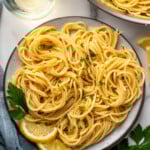 Overhead of plate of lemon pasta garnished with parsley and lemon slices. Glass of white wine, parsley, and lemon pasta surrounding.