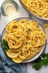 Overhead of plate of lemon pasta garnished with parsley and lemon slices. Glass of white wine, parsley, and lemon pasta surrounding.