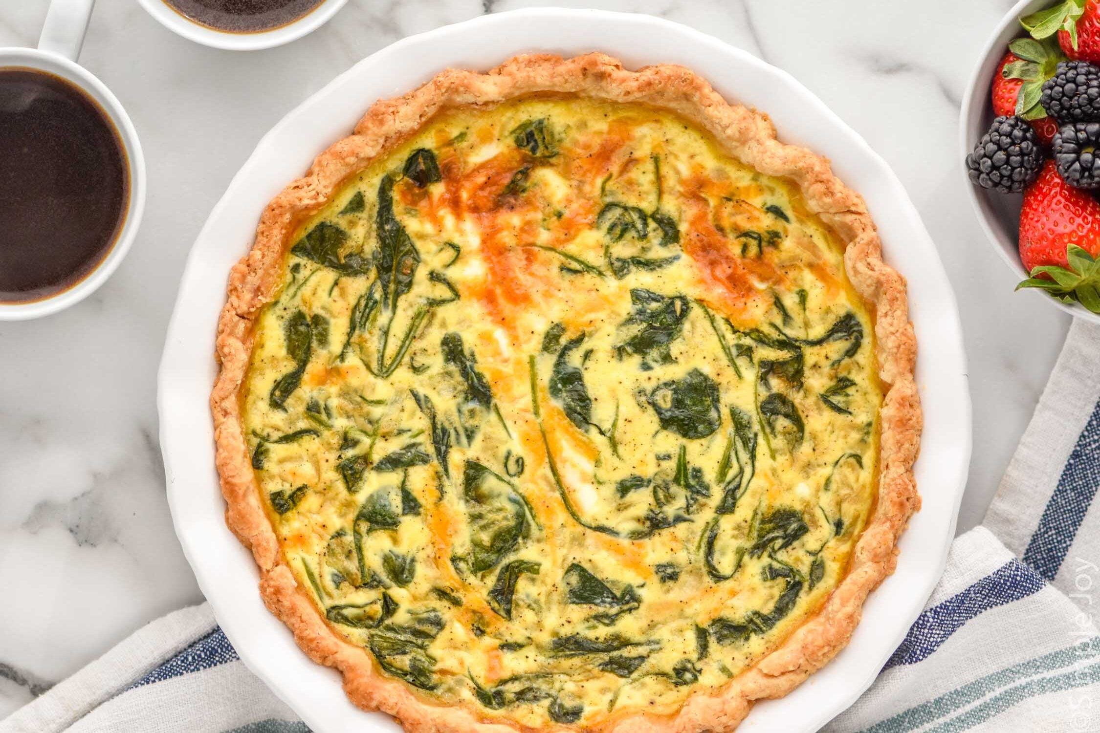 Overhead view of a Spinach Quiche with cups of coffee and bowl of berries beside.