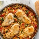 Pinterest graphic for Mediterranean Chicken and Orzo. Says "Mediterranean Chicken & Orzo simplejoy.com" Image shows overhead of skillet of Mediterranean Chicken and Orzo with sliced baguette beside the pan.