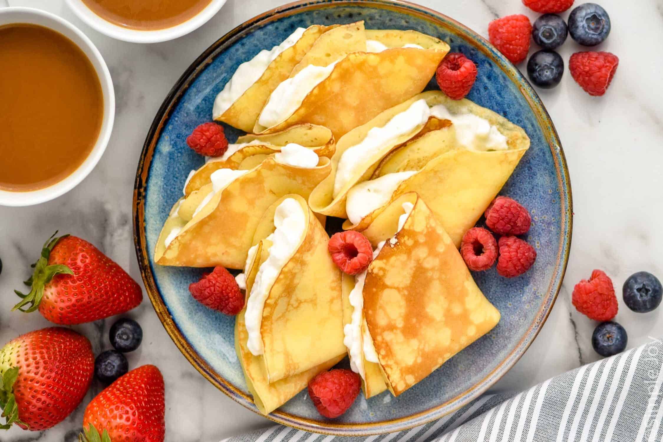 Overhead of plate of crepes stuffed with whipped cream and topped with fresh berries. two mugs of coffee and berries sitting beside plate.