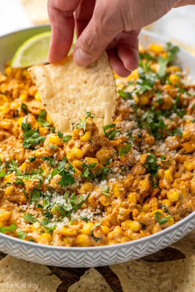 person's hand dipping tortilla chip into bowl of Mexican Street Corn Dip garnished with chopped cilantro