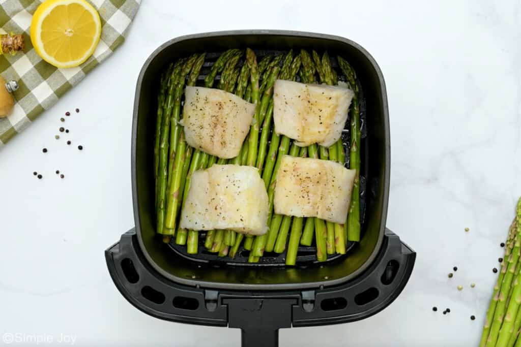 Overhead of air fryer basket of asparagus and cod to make Air Fryer Cod recipe, asparagus and lemon sitting beside
