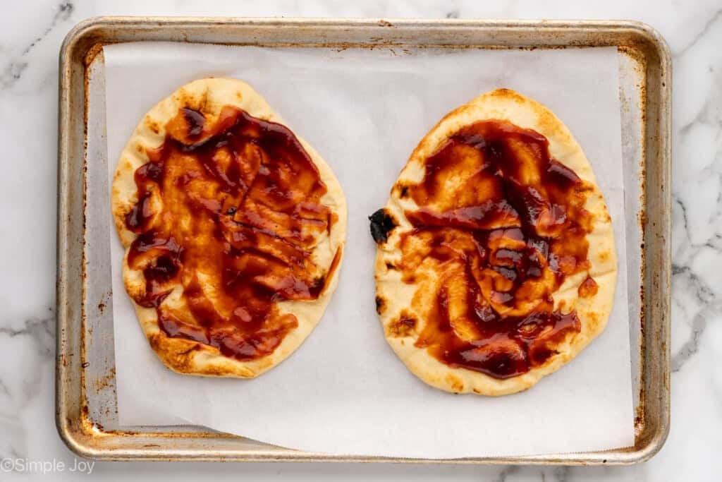 Overhead of baking sheet with two flatbreads topped with bbq sauce to make BBQ Chicken Flatbread Pizza