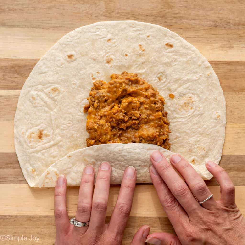 overhead of woman's hands rolling up tortilla of Baked Beef and Cheese Chimichangas ingredients