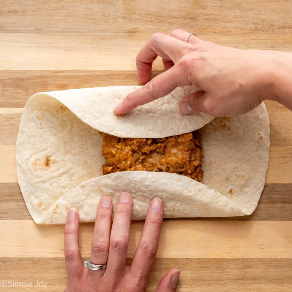 overhead of woman's hands rolling up tortilla of Baked Beef and Cheese Chimichangas ingredients