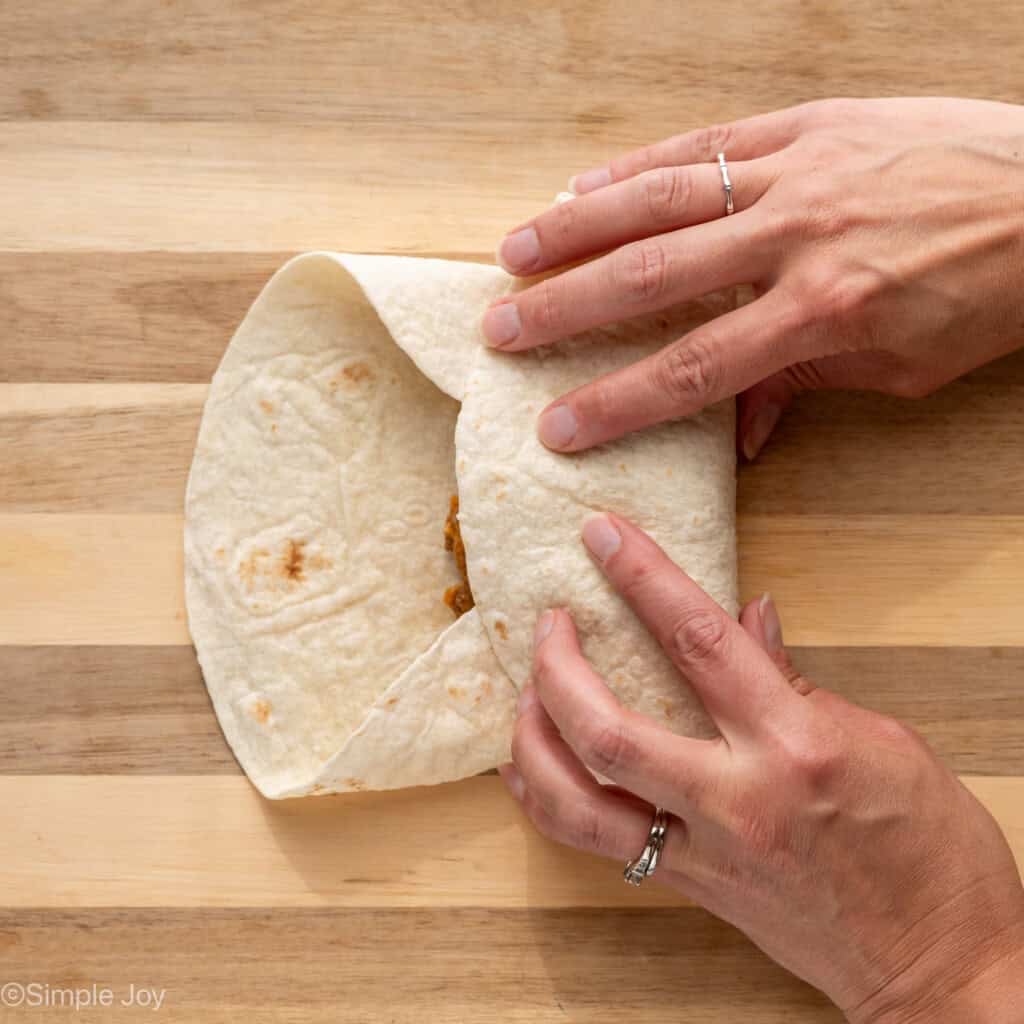 overhead of woman's hands rolling up tortilla of Baked Beef and Cheese Chimichangas ingredients