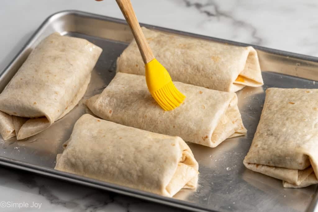 kitchen brushing oil onto Baked Beef and Cheese Chimichangas on a baking sheet