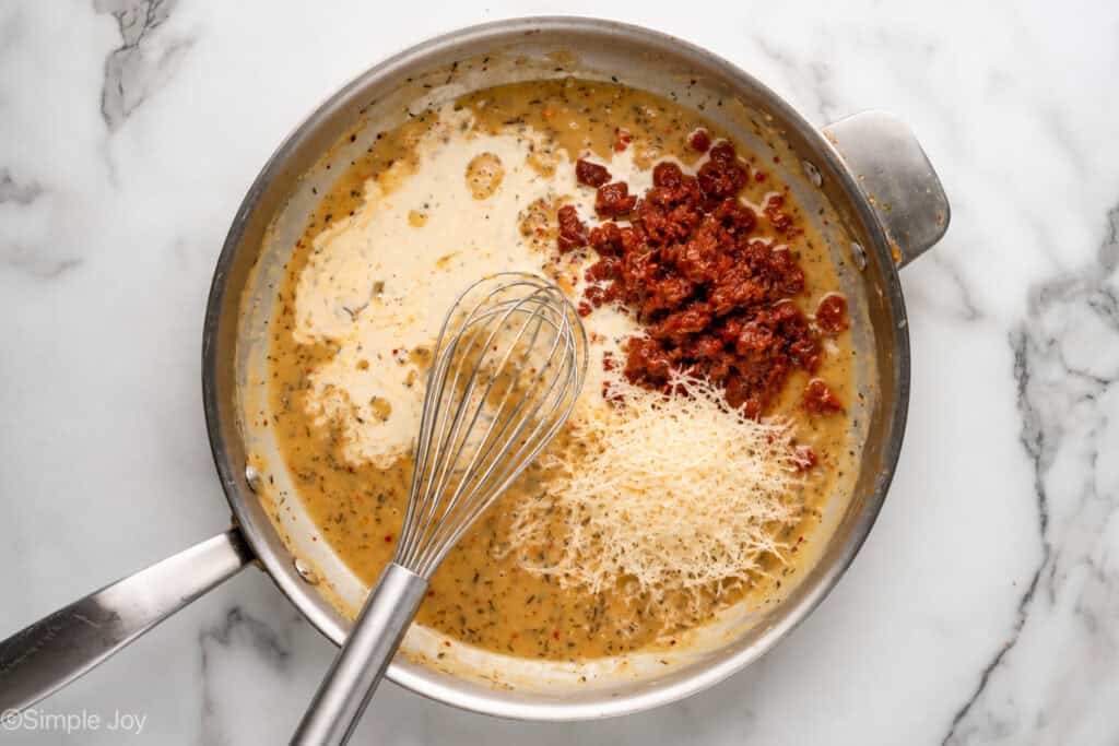 overhead of a skillet with sauce, cream, sun-dried tomatoes, parmesan cheese and a whisk