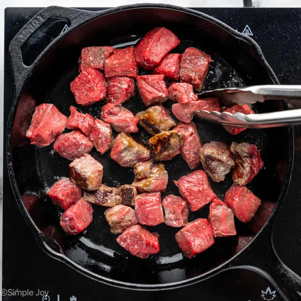 Overhead of skillet of Garlic Butter Steak Bites with tongs to flip them