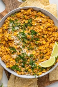 Overhead of bowl of Mexican Street Corn Dip garnished with chopped cilantro and lime wedges, tortilla chips sitting beside