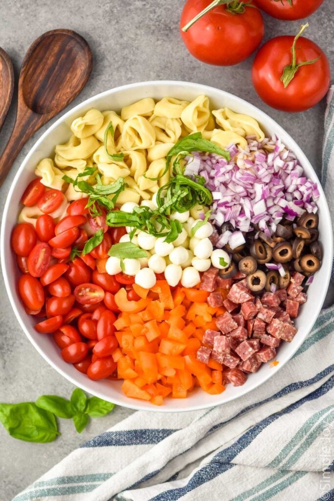Overhead photo of a bowl of ingredients for Tortellini Pasta Salad recipe. Serving spoons and tomatoes beside bowl.