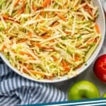 Pinterest graphic for apple slaw. Image shows overhead of bowl of apple slaw with apples and wooden spoons sitting beside. Says "delicious apple slaw simplejoy.com"