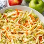 Pinterest graphic for apple slaw recipe. Says "the best apple slaw recipe simplejoy.com" Image shows bowl of apple slaw with apples sitting in background