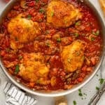 Overhead of skillet of Chicken Cacciatore recipe with tongs, baguette, and two glasses of red wine sitting beside.