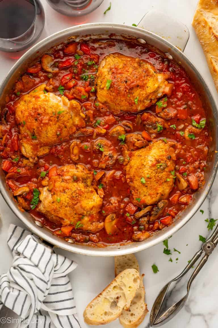 Overhead of skillet of Chicken Cacciatore recipe with tongs, baguette, and two glasses of red wine sitting beside.
