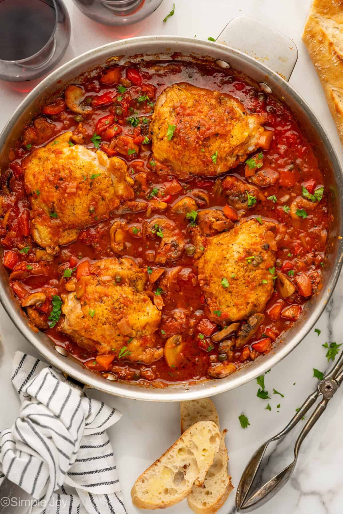 Overhead of skillet of Chicken Cacciatore recipe with tongs, baguette, and two glasses of red wine sitting beside.
