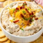 Bowl of crack dip garnished with shredded cheese, bacon, and chives surrounded by crackers.