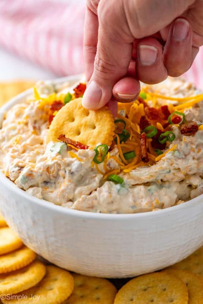 Person's hand dipping cracker into bowl of crack dip garnished with shredded cheese, bacon, and chives.