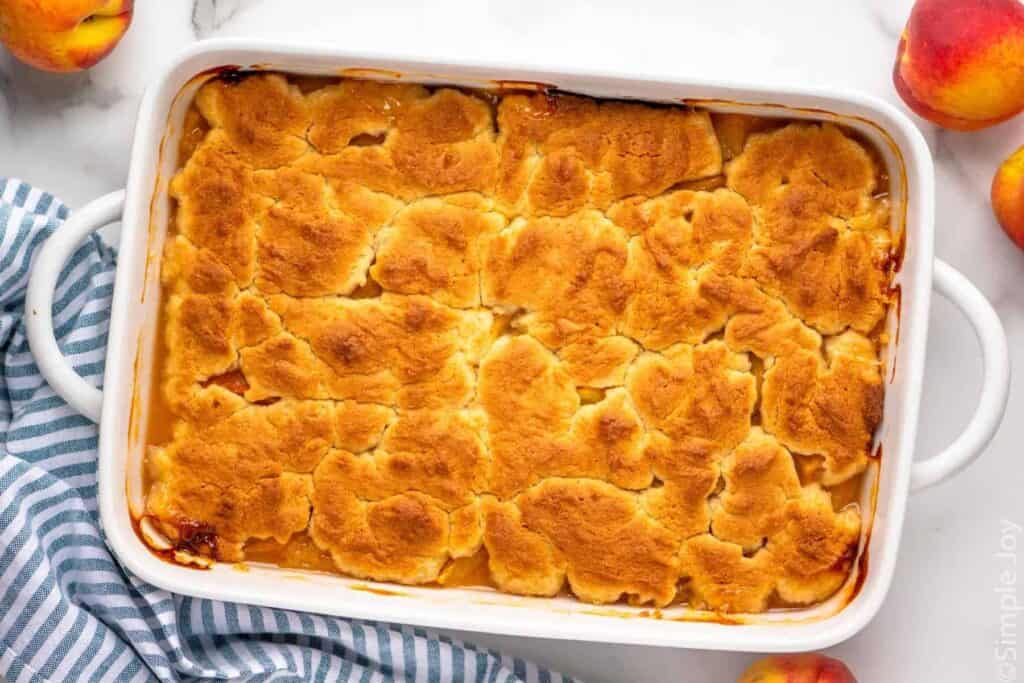 Overhead view of a baking dish of peach cobbler with peaches beside.