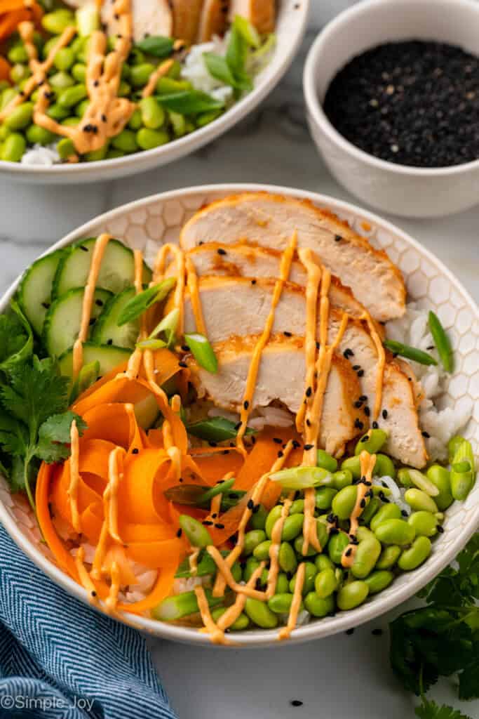 bowl of Honey Sriracha Chicken, slices cucumbers, shredded carrots, and edamame over rice, drizzle of honey sriracha sauce on top