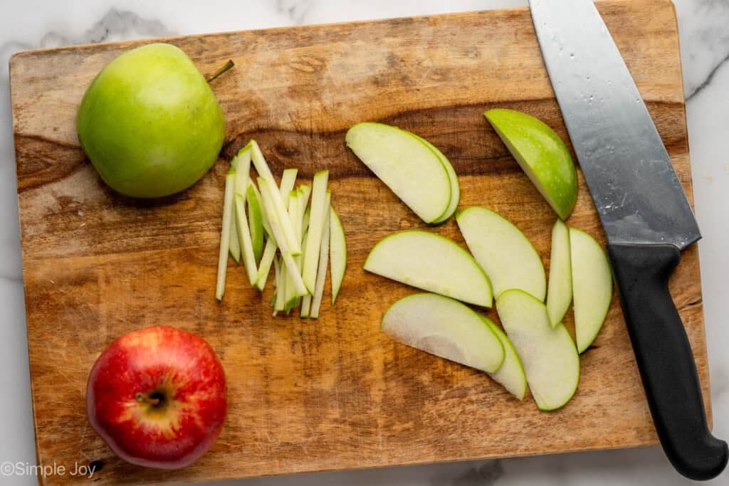 Overhead of apples and apple slices with a knife on a wooden cutting board to make apple slaw