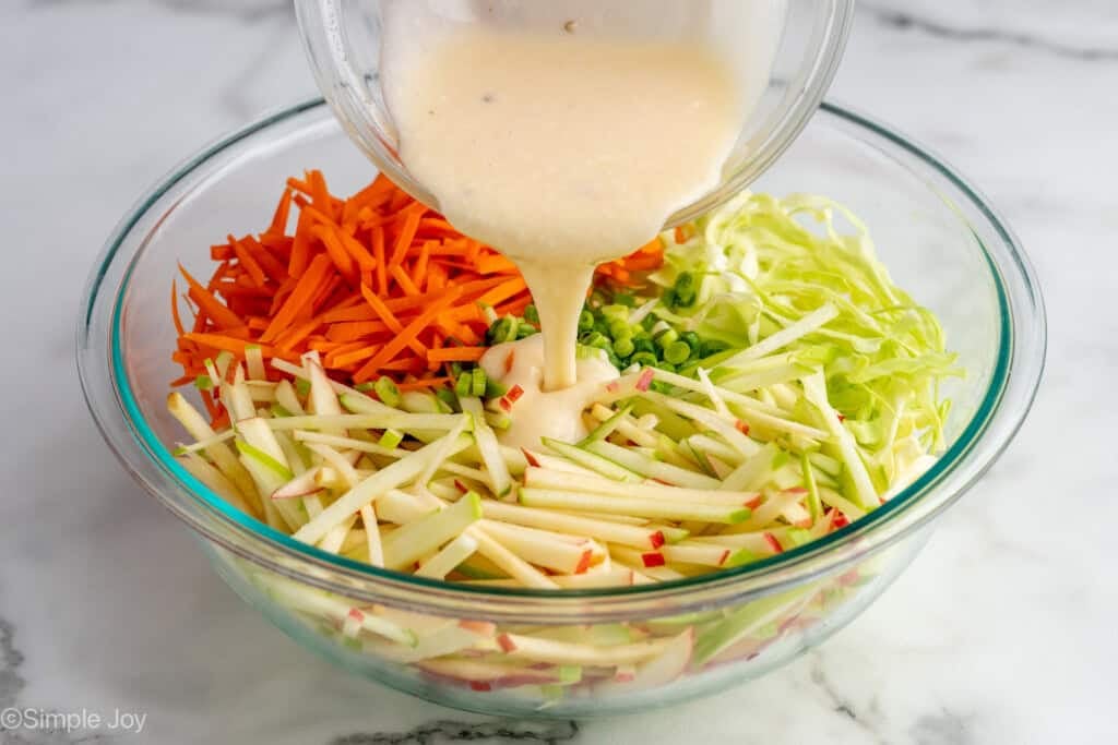 Bowl of apple slaw dressing pouring into glass bowl of apple slaw ingredients