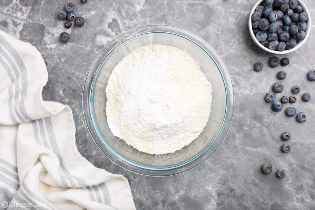 Overhead of glass bowl of blueberry cobbler ingredients with fresh blueberries sitting beside