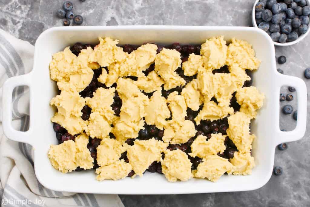 Overhead of baking dish of blueberry cobbler ingredients, fresh blueberries sitting beside.