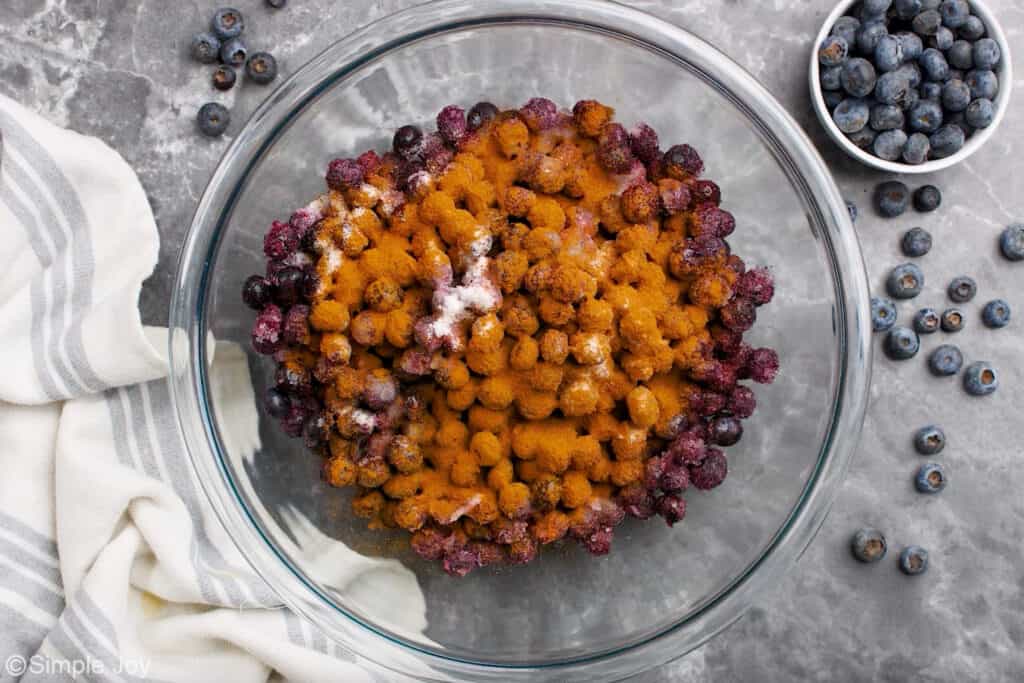 Overhead of large glass bowl of blueberry cobbler ingredients with fresh blueberries sitting beside