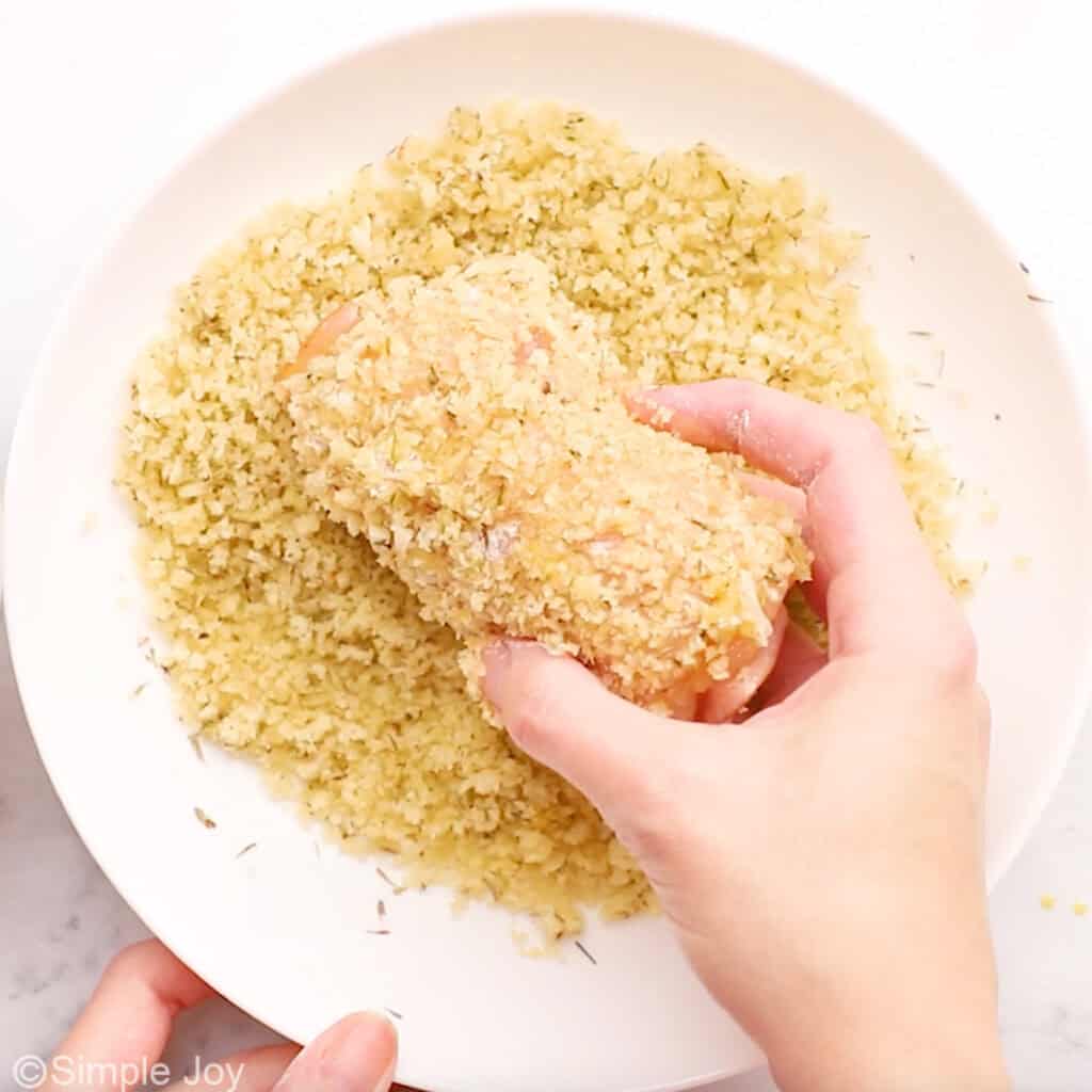 Person's hand holding piece of chicken cordon bleu in plate of breading mixture