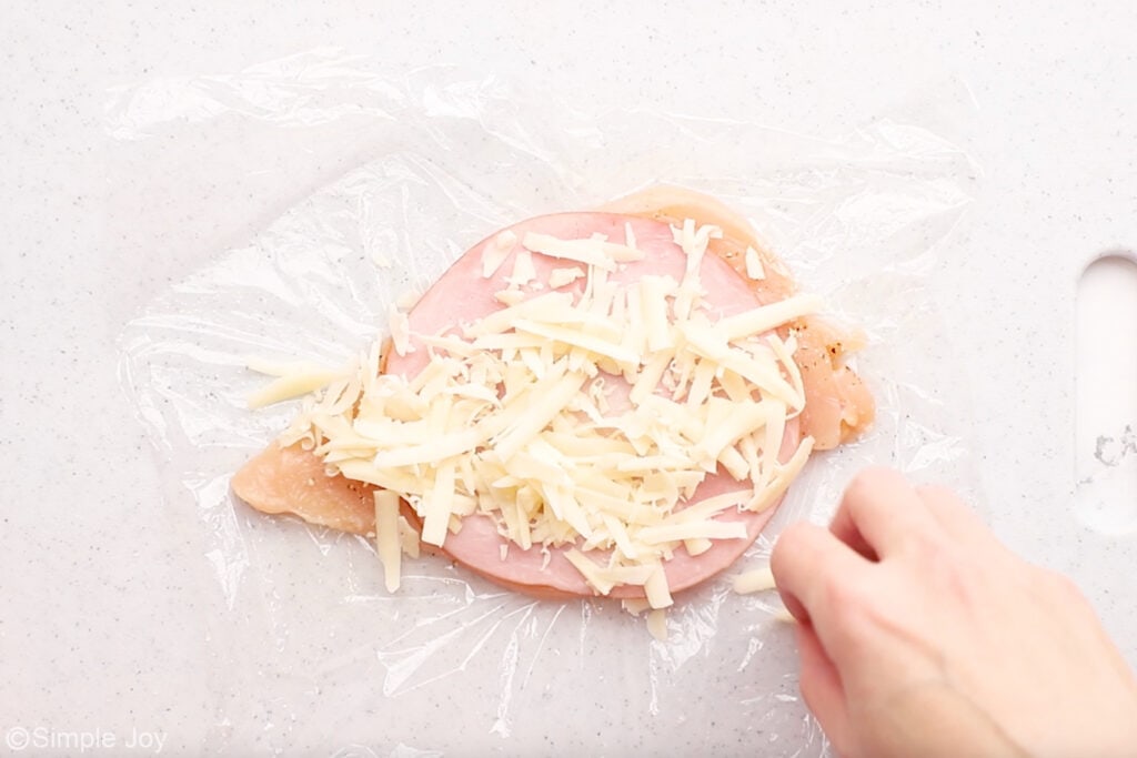 Overhead of chicken cordon bleu ingredients with person's hand sprinkling cheese on top