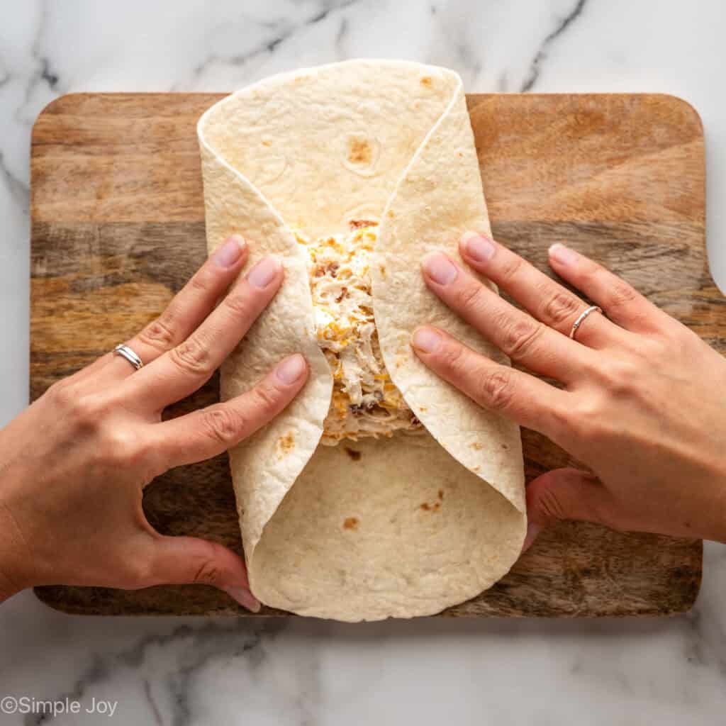 Overhead of person's hands folding tortilla with crack chicken wrap filling ingredients inside