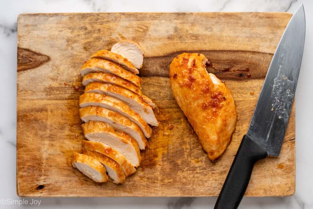 Overhead of Honey Sriracha Chicken and large knife on a wooden cutting board to make Honey Sriracha Chicken Bowls