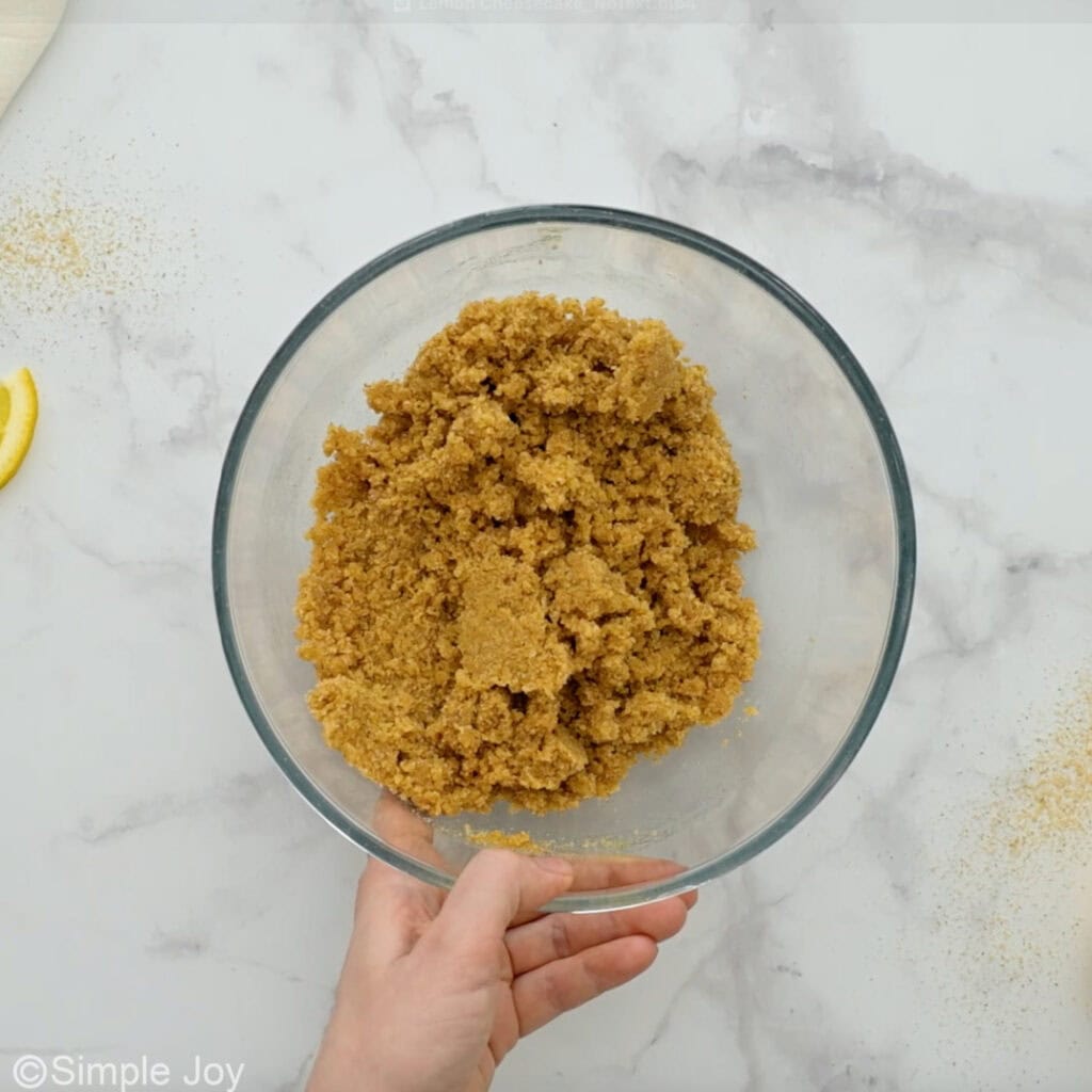 Overhead of person's hand holding glass bowl of lemon cheesecake crust ingredients
