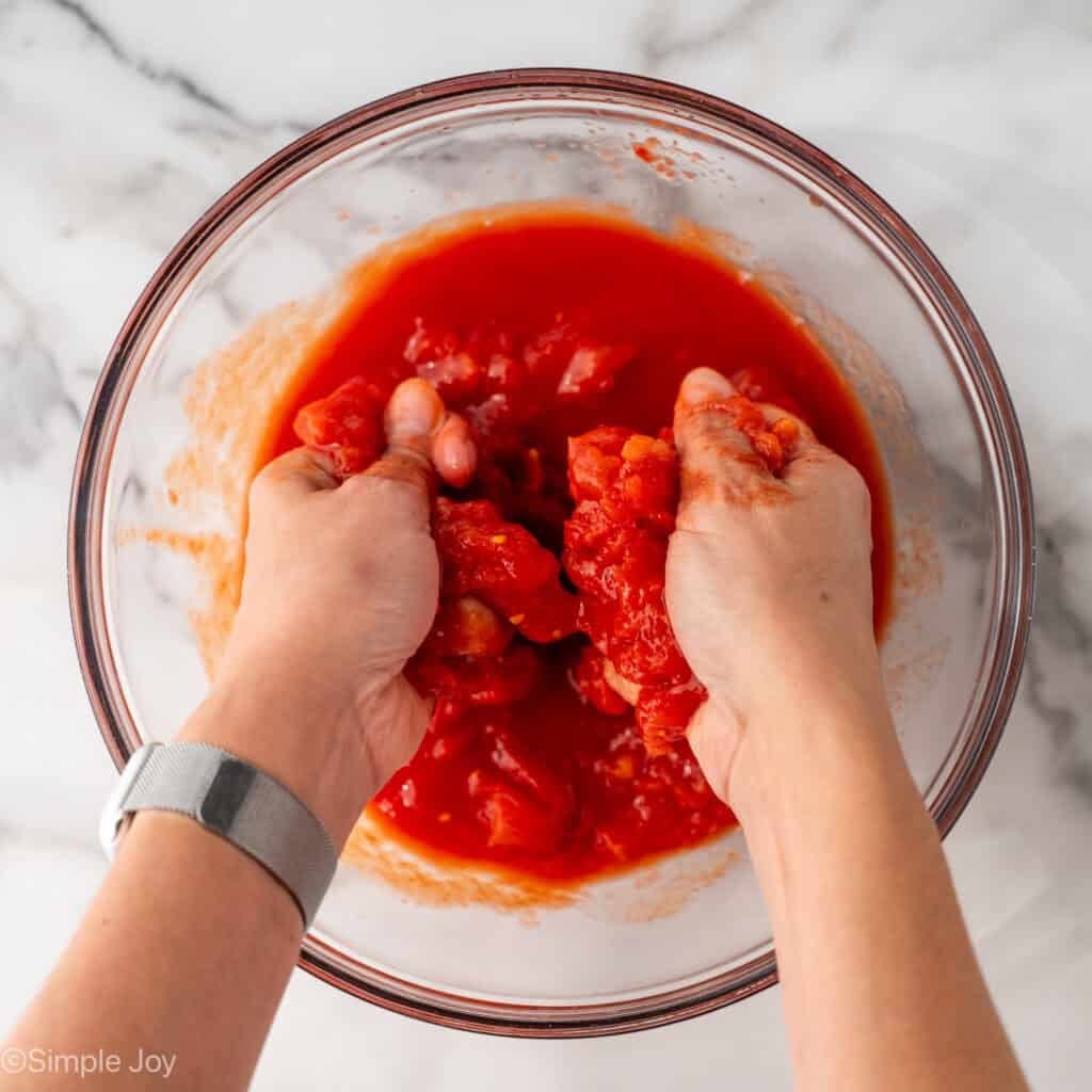 Overhead of person's hands squeezing tomatoes in a glass bowl to make Pasta Puttanesca