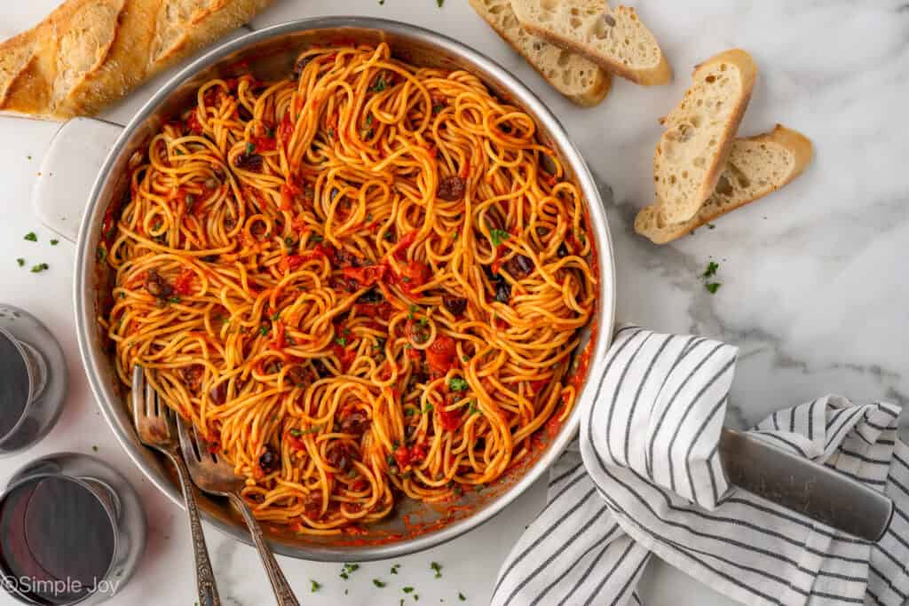 Overhead of skillet of Pasta Puttanesca with two glasses of red wine and sliced baguette sitting beside