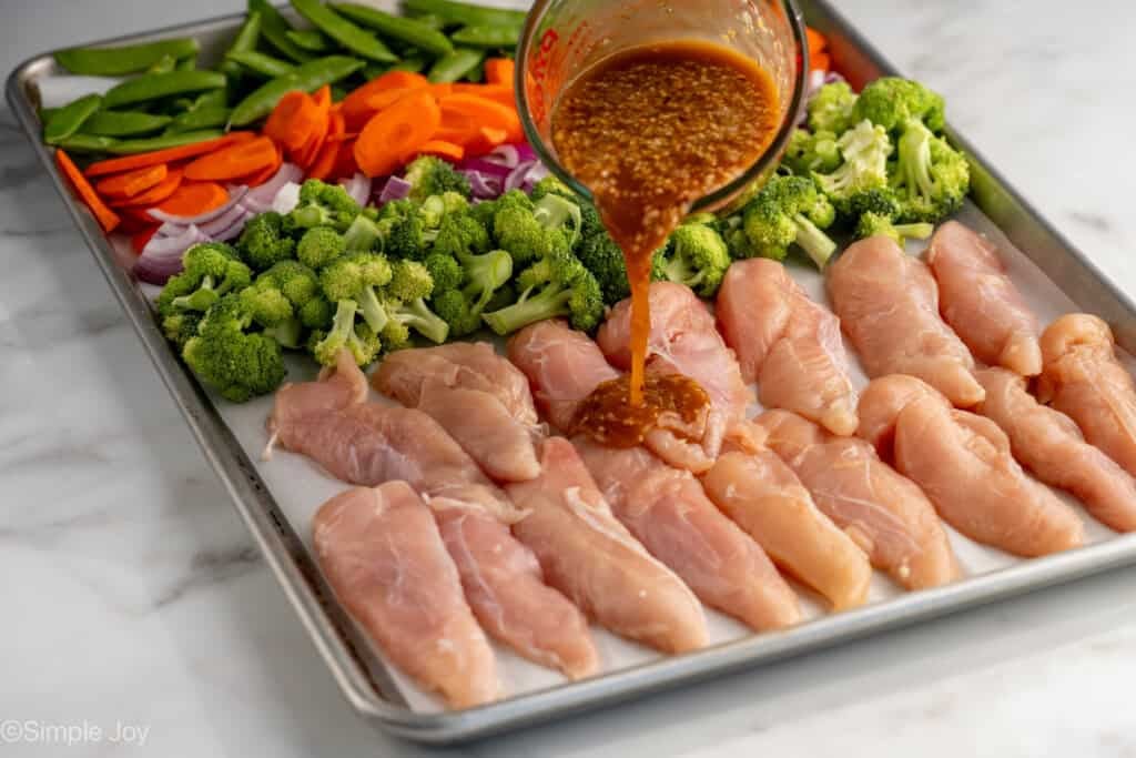 Glass measuring cup of Honey Garlic Chicken and Vegetables ingredients pouring onto chicken and vegetables on a lined baking sheet to make Honey Garlic Chicken and Vegetables