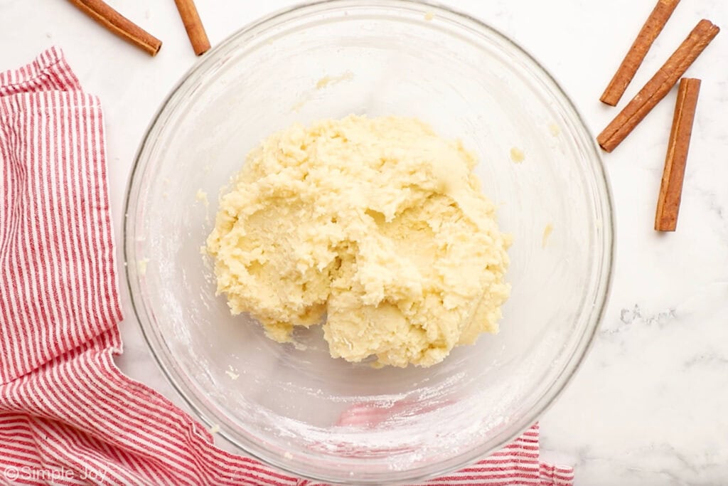 Overhead of glass bowl of snickerdoodle ingredients with cinnamon sticks sitting beside