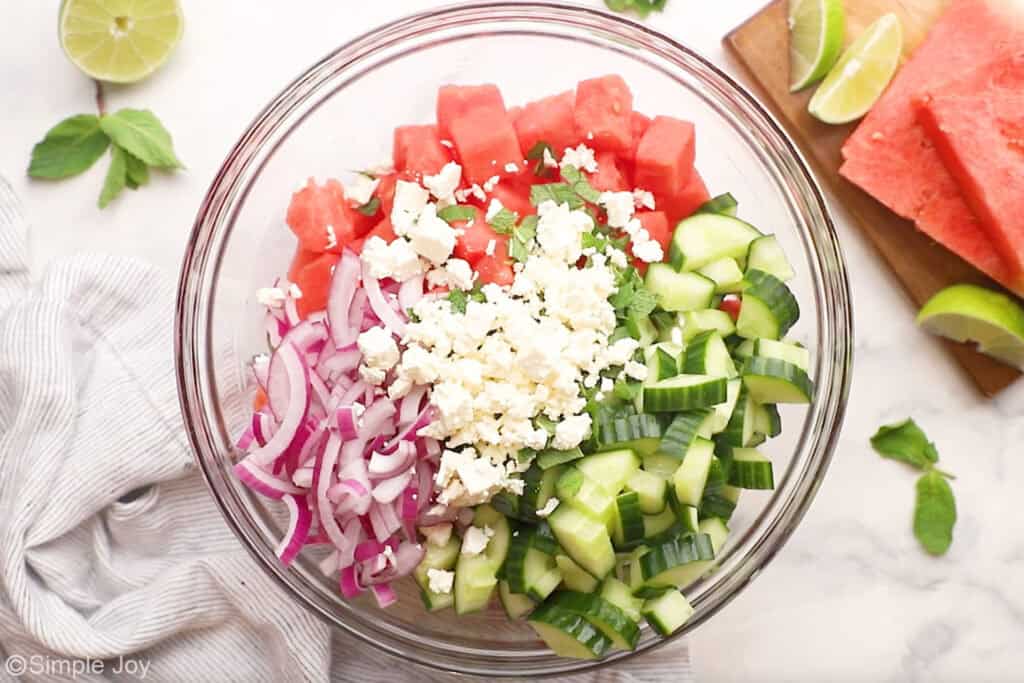 Overhead of glass bowl of watermelon salad ingredients with fresh mint leaves, lime wedges, and watermelon sitting beside