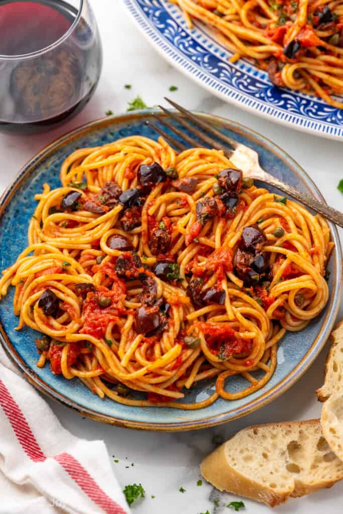 Plate of Pasta Puttanesca with fork, glass of red wine and sliced baguette sitting beside