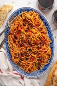Overhead of platter of Pasta Puttanesca with two forks, glass of red wine and sliced baguette sitting beside